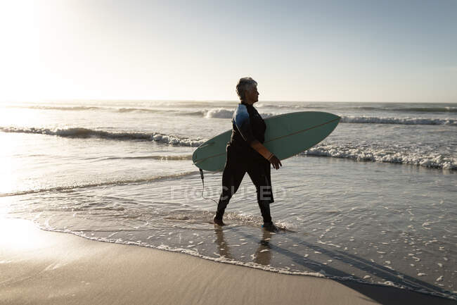 Mulher americana africana sênior carregando prancha de surf andando na praia. viagem férias aposentadoria estilo de vida conceito — Fotografia de Stock