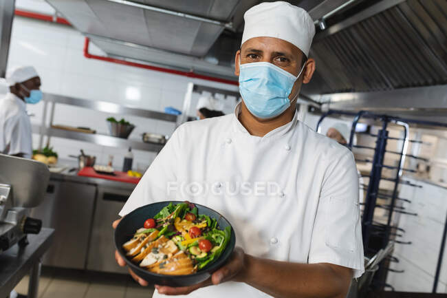Porträt eines männlichen Profikochs gemischter Rasse mit fertigem Gericht, der Gesichtsmaske trägt. Arbeit in einer belebten Restaurantküche während der Coronavirus-Pandemie 19. — Stockfoto