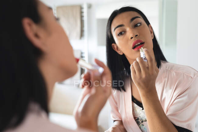 Transgender-Frau mit gemischter Rasse schaut in Badezimmerspiegel und setzt Lippenstift auf. Isolationshaft während der Quarantäne. — Stockfoto