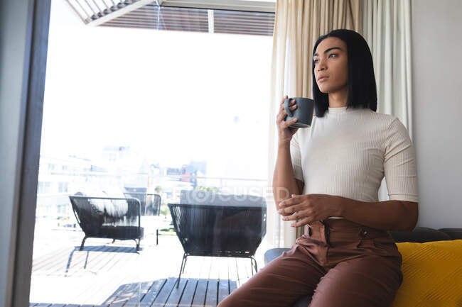 Transgender-Frau mit gemischter Rasse sitzt im Wohnzimmer und schaut aus dem Fenster mit einer Tasse Kaffee. Isolationshaft während der Quarantäne. — Stockfoto
