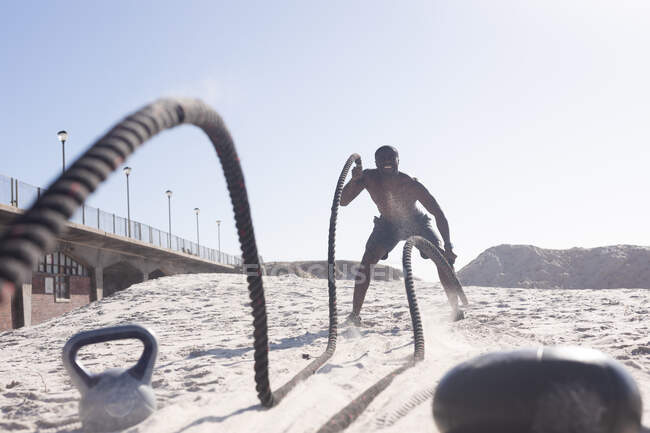 African american man exercising with rope and kettle on beach. healthy outdoor lifestyle fitness training. — Fotografia de Stock