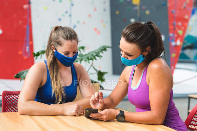 Две счастливые кавказки в масках с помощью смартфона у стены для скалолазания. фитнес и досуг в тренажерном зале во время коронавируса ковид 19 пандемии. — стоковое фото