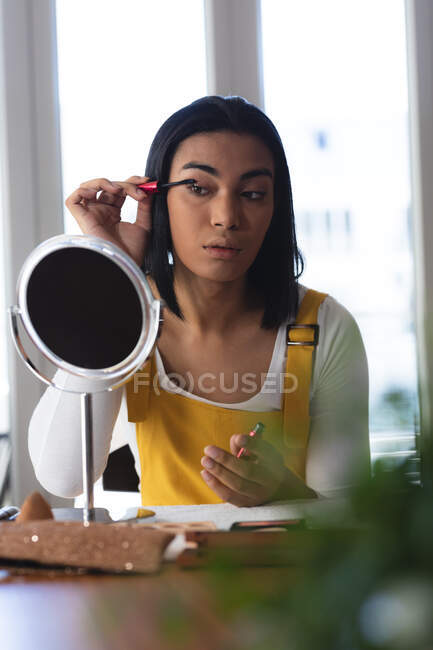 Mixed race transgender woman sitting at table looking in mirror putting on mascara. staying at home in isolation during quarantine lockdown. — Stock Photo