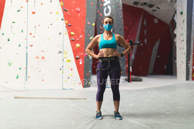 Porträt einer Kaukasierin mit Gesichtsmaske, die an einer Indoor-Kletterwand steht. Fitness und Freizeit im Fitnessstudio während der Coronavirus-Pandemie 19. — Stockfoto