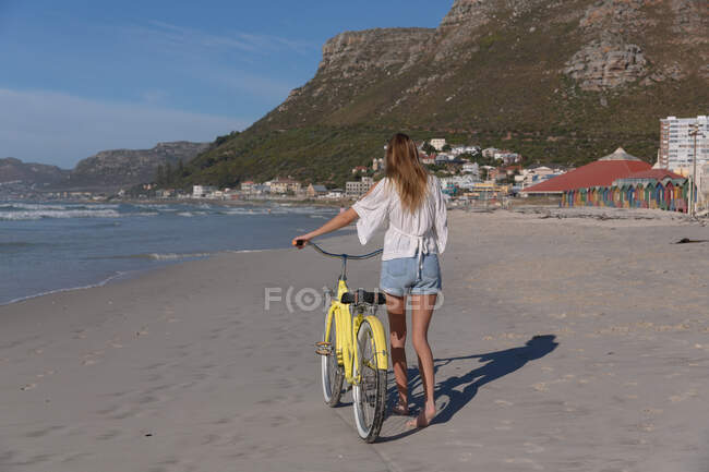 Rear view of caucasian woman in white top and shorts walking with a bicycle at the beach. healthy outdoor leisure time by the sea. - foto de stock