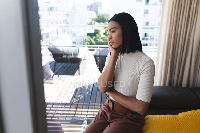 Mujer transgénero de raza mixta sentada en el pensamiento en la sala de estar en el día soleado. permanecer en casa aislado durante el bloqueo de cuarentena. - foto de stock