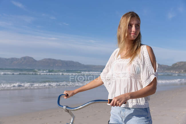 Mujer caucásica caminando y llevando una bicicleta en la playa. tiempo de ocio al aire libre saludable junto al mar. - foto de stock