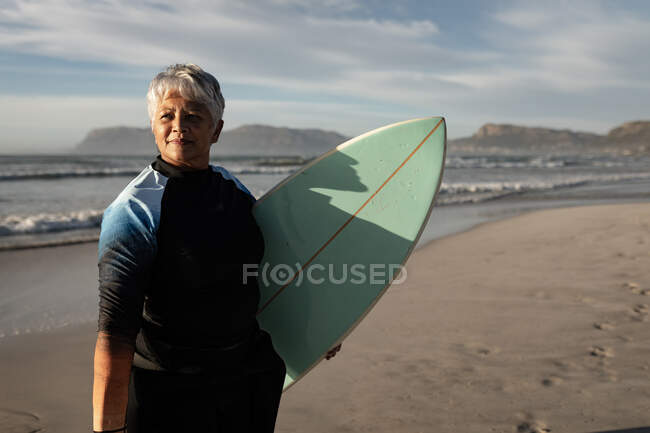 Mujer afroamericana mayor sosteniendo tabla de surf de pie en la playa. viaje vacaciones retiro estilo de vida concepto - foto de stock