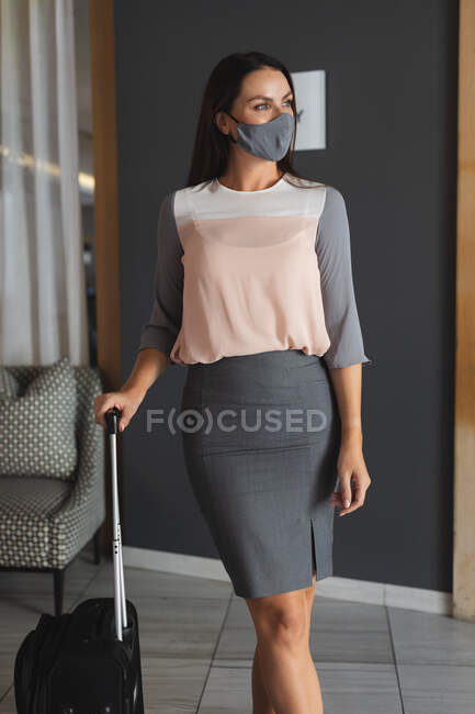 Портрет белой женщины в маске, стоящей в вестибюле гостиницы с чемоданом. отель для деловых поездок во время пандемии коронавируса. — стоковое фото