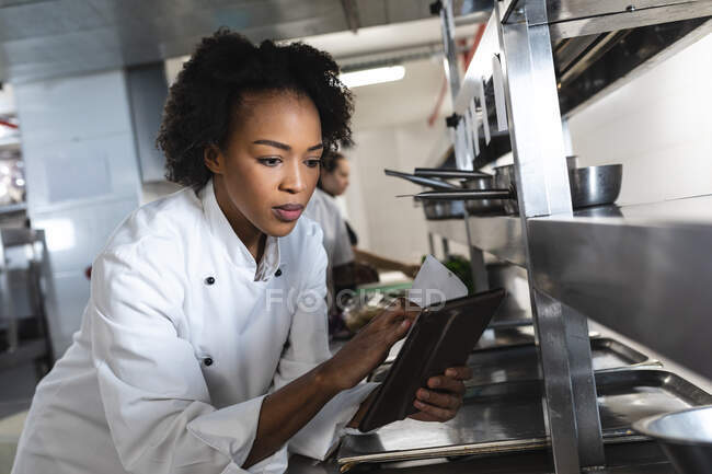 Mixed race professional chef looking at tablet with colleague in background. working in a busy restaurant kitchen. — Stock Photo