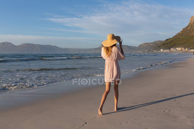 Белая женщина в пляжном покрывале и шляпе, развлекающаяся прогулкой по пляжу. здоровый отдых на открытом воздухе у моря. — стоковое фото
