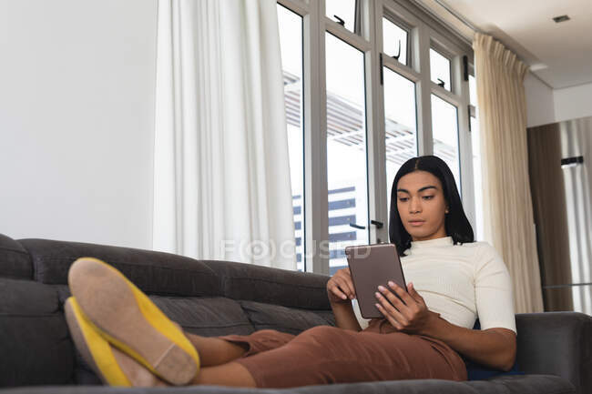 Transgender-Frau mit gemischter Rasse entspannt sich im Wohnzimmer auf dem Sofa liegend mit Tablet. Isolationshaft während der Quarantäne. — Stockfoto