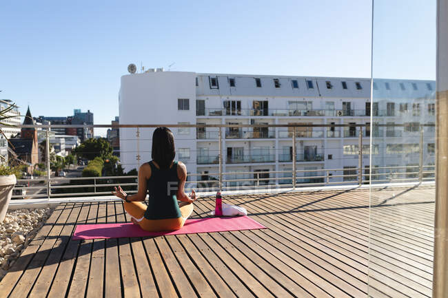 Задний вид смешанной расы трансгендерных женщин практикующих йогу медитации на террасе на крыше на солнце. оставаться дома в изоляции во время карантинной изоляции. — стоковое фото