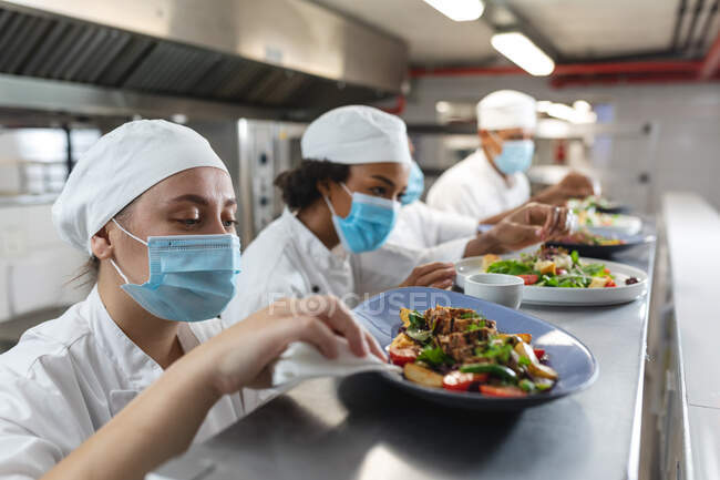 Diverse männliche und weibliche Profiköche verschenken Gerichte mit Mundschutz. Arbeit in einer belebten Restaurantküche während der Coronavirus-Pandemie 19. — Stockfoto