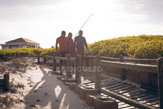 Père afro-américain et ses deux fils avec des cannes à pêche marchant ensemble sur le pont. vacances à la plage d'été et concept loisirs. — Photo de stock