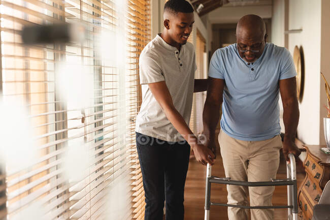Joven afroamericano ayudando a su padre a caminar con el marco de caminar en casa. concepto de amor y cuidado de ancianos - foto de stock
