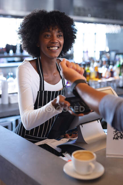 Barista afroamericana sonriente vistiendo delantal, usando terminal de pago. café independiente, pequeña empresa exitosa. - foto de stock