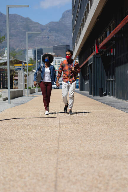 Мужчина смешанной расы и африканская американка в масках, ходят, держатся за руки. тусоваться вместе во время пандемии коронавируса 19. — стоковое фото