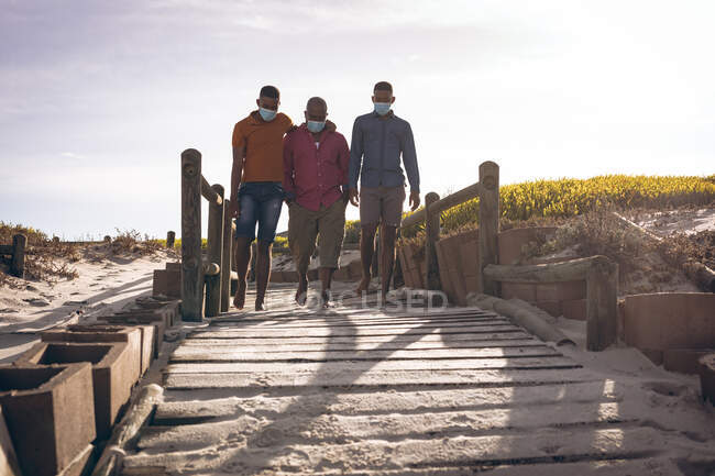 Африканский отец-американец и два его сына в масках для лица идут вместе по мосту. правила летнего отдыха на пляже во время пандемии ковид-19. — стоковое фото
