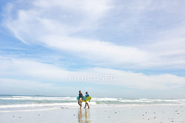 Afroamericano padre e hijo con tablas de surf caminando hacia las olas en la playa. vacaciones de playa de verano y concepto de ocio. - foto de stock