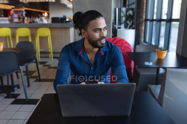 Smiling mixed race man sitting, using smartphone and laptop in cafe. digital creatives on the go. — Stock Photo