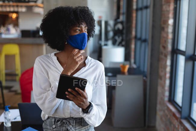 Donna afroamericana indossando maschera facciale e utilizzando tablet in caffè. creativi digitali in movimento durante coronavirus covid 19 pandemia. — Foto stock