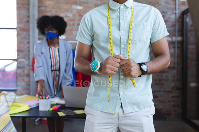 Midsection of mixed race designer wearing tailor's meter, african american female in background. independent creative fashion design business during coronavirus covid 19 pandemic — Stock Photo