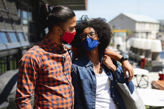 Hombre de raza mixta y mujer afroamericana usando máscaras, abrazando. pasando el rato juntos durante la pandemia del coronavirus covid 19. - foto de stock