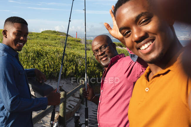 Der afroamerikanische Vater und seine beiden Söhne lächeln zusammen, während sie ein Selfie auf der Brücke machen. Sommer-Strandurlaub und Freizeitkonzept. — Stockfoto