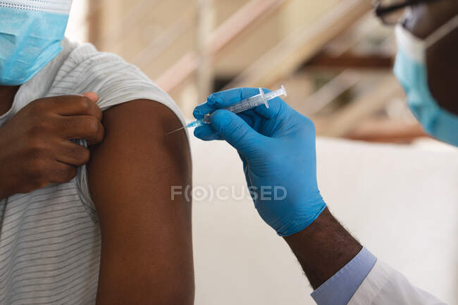 Médico afroamericano sénior inyectando la vacuna covid-19 en el hombre afroamericano en casa. vacunación para la prevención del concepto de brote de coronavirus - foto de stock