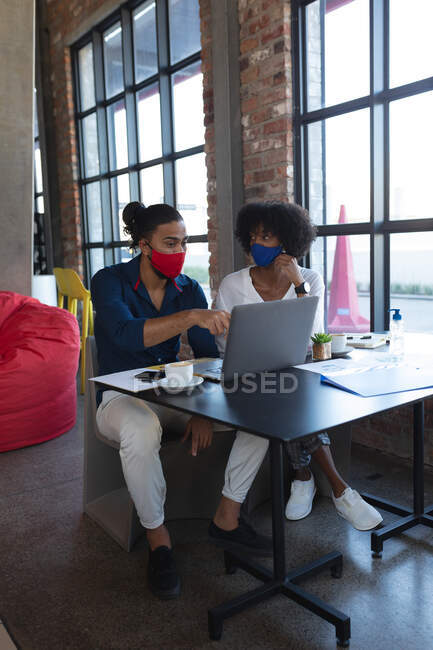 African american woman and mixed race man wearing face masks sitting in cafe using laptop. digital creatives on the go during coronavirus covid 19 pandemic. — Stock Photo