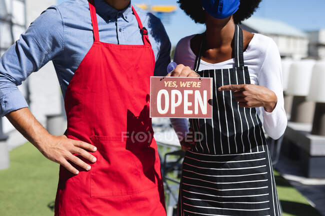 Midsection of diverse male and female waiters wearing face masks holding open sign. independent cafe, business during coronavirus covid 19 pandemic. — Stock Photo