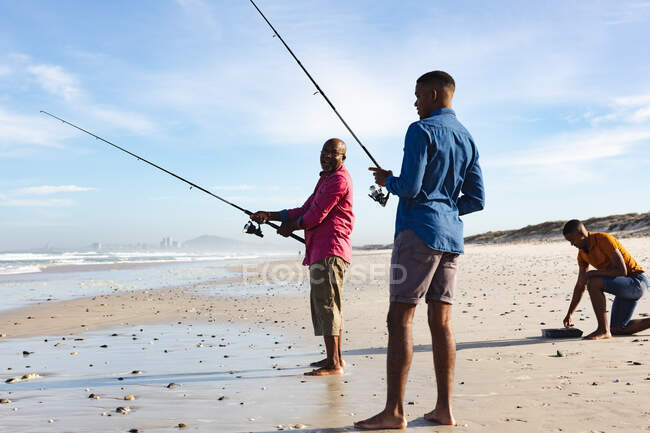 Afrikanischer Vater und seine beiden Söhne mit Angelruten beim gemeinsamen Angeln am Strand. Sommer-Strandurlaub und Freizeitkonzept. — Stockfoto