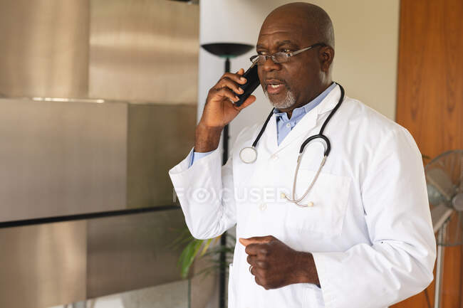 African american senior male doctor talking on smartphone at home. distant communication and telemedicine consultation concept. — Stock Photo