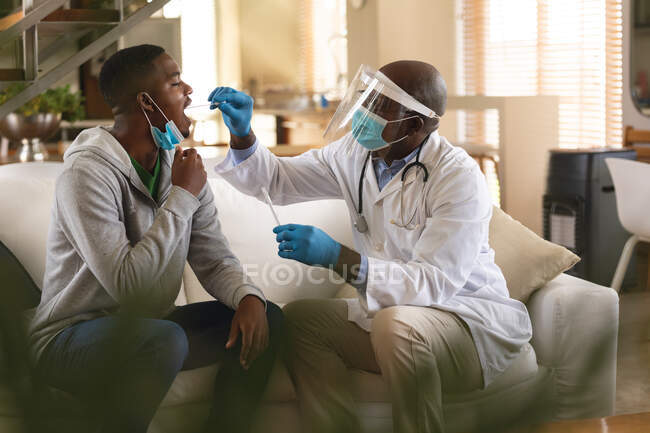 Médico varón sénior afroamericano realizando una prueba de frotis de garganta en el hombre afroamericano en casa. pruebas médicas para la prevención del concepto de brote de coronavirus - foto de stock