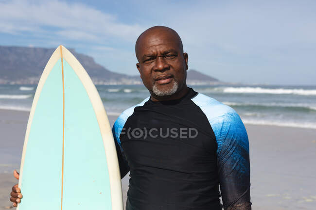 Портрет афроамериканського старшого чоловіка з дошкою для серфінгу, що стоїть на пляжі. літній пляжний відпочинок і концепція дозвілля . — стокове фото