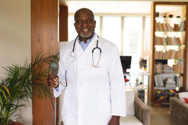 Portrait of african american senior male doctor holding glasses while standing at home. distant communication and telemedicine consultation concept. — Stock Photo