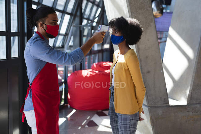 Hombre de raza mixta que usa mascarilla facial y comprueba la temperatura de la mujer afroamericana. café independiente, negocio durante coronavirus covid 19 pandemia. - foto de stock