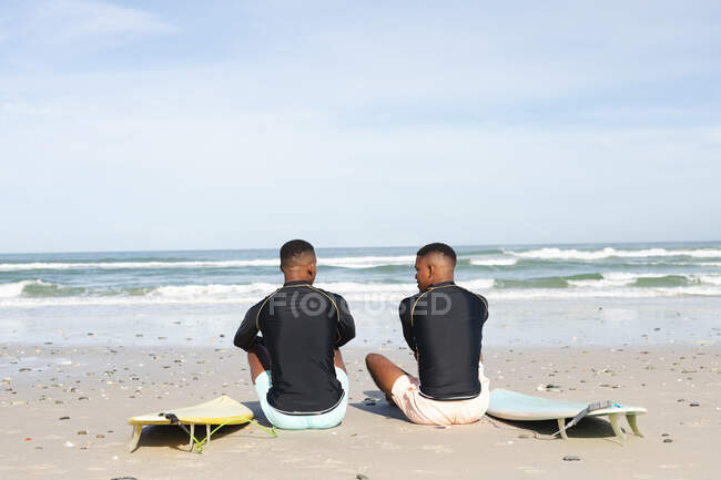 Rear view of african american brothers with surfboards sitting at the beach. summer beach holiday and leisure concept. — Stock Photo
