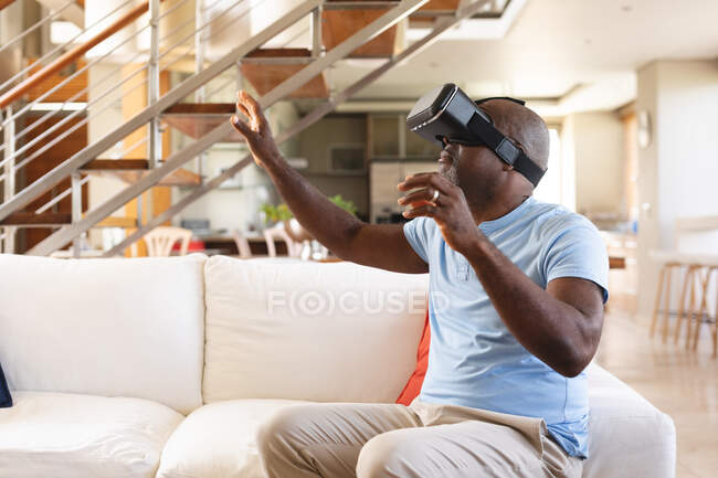 Uomo anziano afroamericano gesticolare mentre indossa auricolare vr seduto sul divano a casa. concetto di tecnologia di intrattenimento — Foto stock