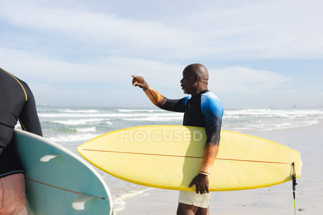Hombre mayor afroamericano con tabla de surf apuntando hacia las olas en la playa. vacaciones de playa de verano y concepto de ocio. - foto de stock