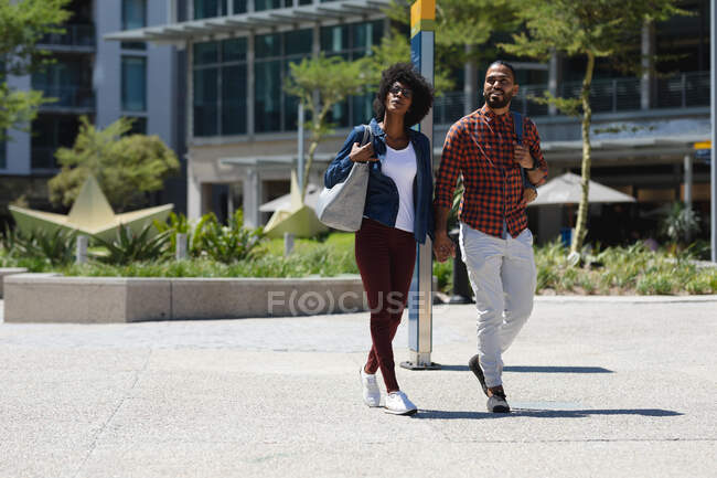 Hombre de raza mixta y mujer afroamericana caminando, tomados de la mano. pasando el rato juntos en un día soleado. - foto de stock