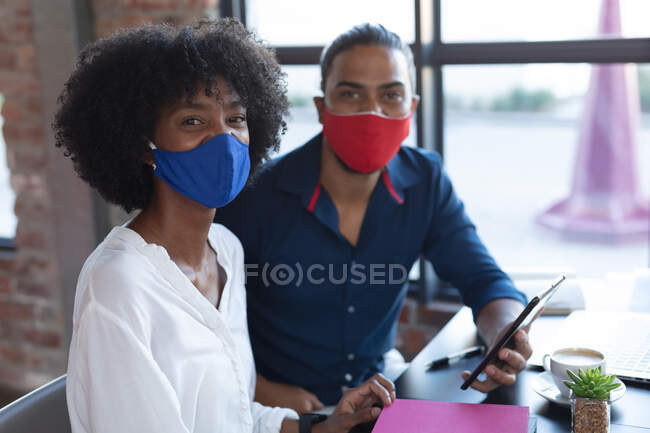 Smiling mixed race man and african american woman wearing face mask sitting, using tablet in cafe. digital creatives on the go during coronavirus covid 19 pandemic. — Stock Photo