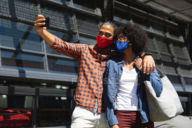 Мужчина смешанной расы и африканская американка в масках, обнимаются, делают селфи. тусоваться вместе во время пандемии коронавируса 19. — стоковое фото