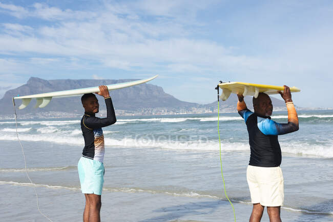 Portrait of african american father and son carrying surfboards on their heads at the beach. summer beach holiday and leisure concept. — Stock Photo