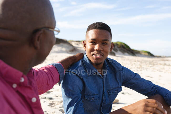 African american father talking to his son while sitting at the beach. summer beach holiday and leisure concept. — Stock Photo