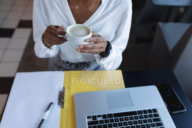 Midsection of african american woman drinking coffee, using laptop and working in cafe. digital creatives on the go. — Stock Photo