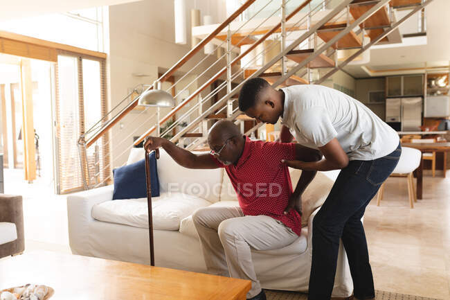 African american young man helping his father with back pain to get up from the couch at home. elderly love and care concept — Stock Photo