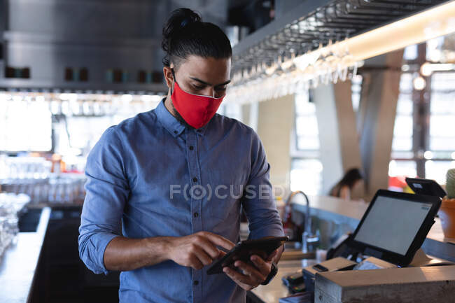 Mixed race male barista wearing face mask, using tablet. independent cafe, business during coronavirus covid 19 pandemic. — Stock Photo
