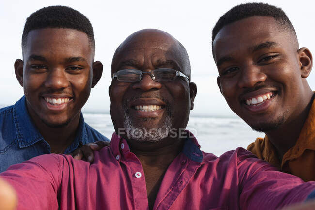 Retrato del padre afroamericano y sus dos hijos tomando un selfie en la playa. vacaciones de playa de verano y concepto de ocio. - foto de stock